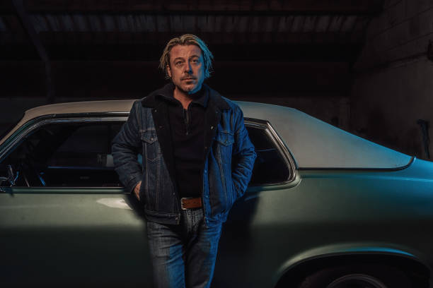 Man with blond hair in a jeans jacket stands by an American classic muscle car.